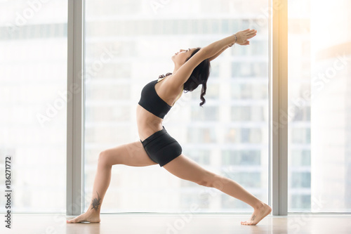 Young sporty woman practicing yoga, standing in Warrior I exercise, Virabhadrasana one pose, working out, wearing sportswear, black tank top, shorts, full length, near floor window with city view