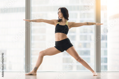 Young attractive woman practicing yoga, standing in Warrior Two exercise, Virabhadrasana II pose, working out, wearing sportswear, black tank top, shorts, full length, near floor window with city view