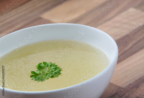 Rich chicken broth with green parsley on top. White bowl of chicken bouillon on wooden background. Closeup view.