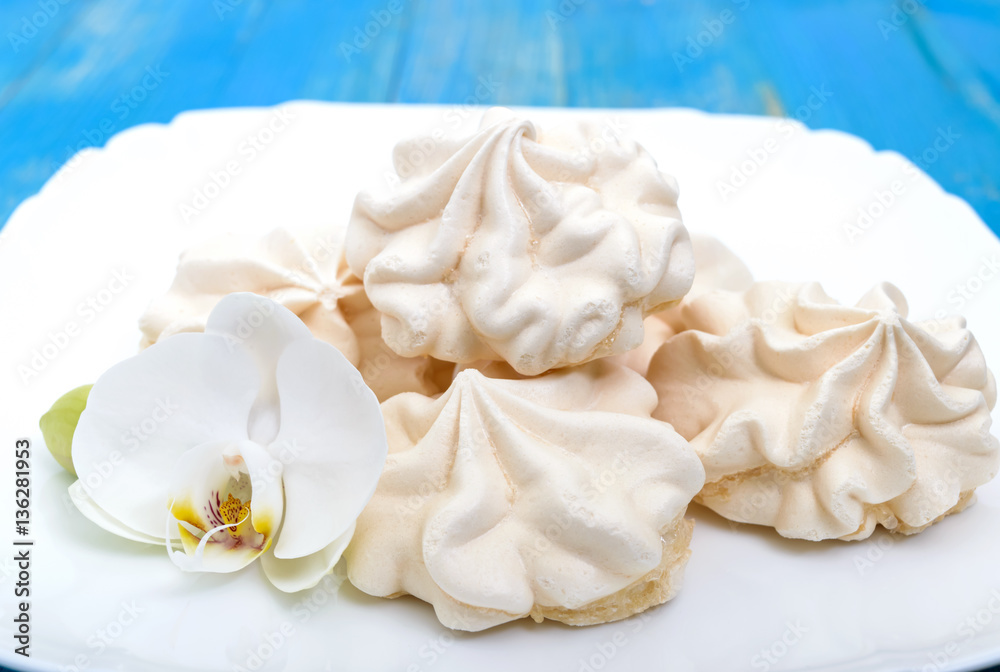 Light air vanilla meringue on a white plate with orchid flower on a blue background. Close-up.