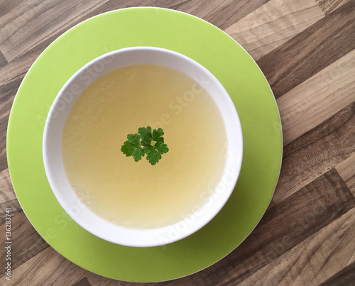 Chicken broth with green parsley on top. White bowl of chicken bouillon on wooden background. Top view.