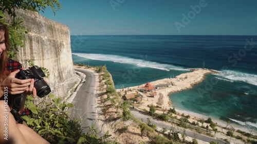 Young caucaasian woman taking pictures sitting on edge of a Melasti beach cliff overlooking roads and turquoise ocean on a sunny summer day in Bali, Indonesia. Shot with sony a7s on a slider photo