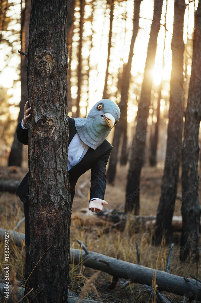 Creepy man in a suit and a rubber bird mask, standing in the sunset forest, behind the tree, waving to the camera Creepy man in a suit and a rubber bird mask, standing in the sunset forest, behind the tree, waving to the camera
