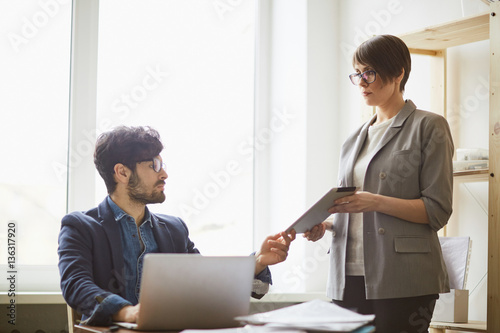 Young boss at workplace: successful middle eastern man looking up from laptop to check schedule at tablet in hands of his female colleague standing by him