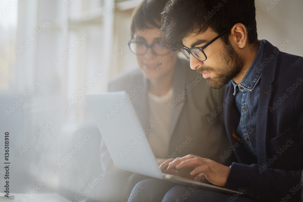 Portrait of two creative business people working together, focus on young middle eastern man wearing beard and glasses busy typing while woman sitting beside him assisting and giving instructions