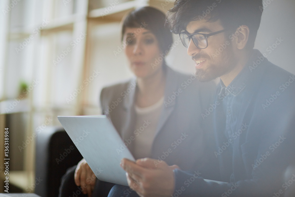 Young businessman wearing creative haircut and glasses smiling while looking at laptop screen and discussing project with colleague Young businessman wearing creative haircut and glasses smiling while looking at laptop screen and discussing project with colleague