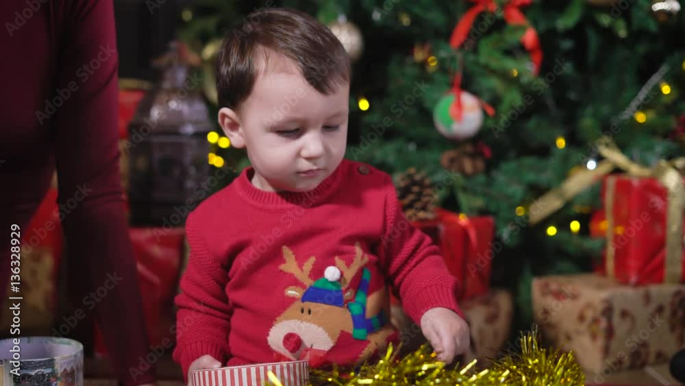 The kid at the Christmas tree. Interested baby playing with a gift box. The kid saw a golden tinsel and pushed her foot. The boy has a good mood, because all around it and reminds us of Christmas. The kid at the Christmas tree. Interested baby playing with a gift box. The kid saw a golden tinsel and pushed her foot. The boy has a good mood, because all around it and reminds us of Christmas.