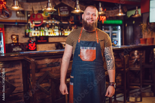 Bearded barman with tattoos and watches wearing an apron standing near the bar.