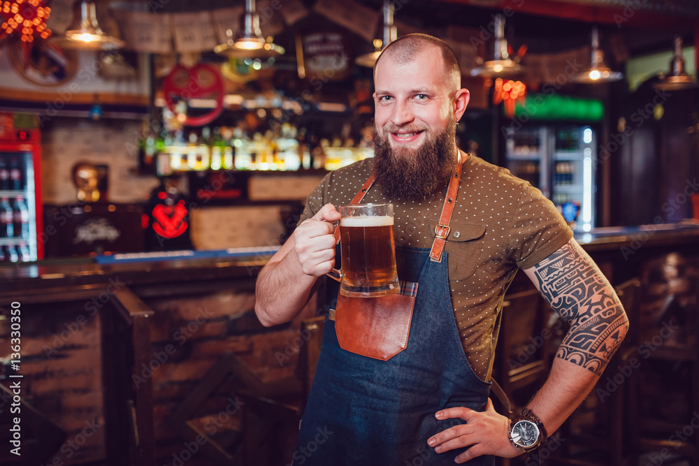 Bearded barman with tattoos wearing an apron standing near the bar and holding a glass of beer