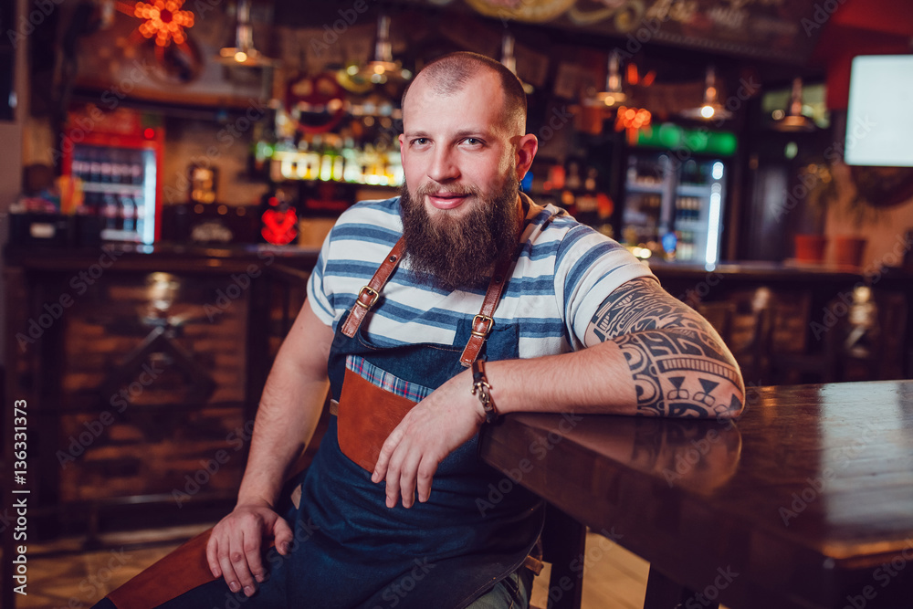 Bearded barman with tattoos wearing an apron sitting at the bar. Bearded barman with tattoos wearing an apron sitting at the bar.