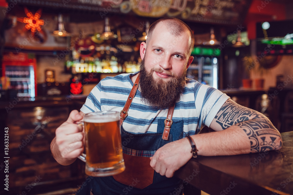 Bearded barman with tattoos wearing an apron sitting at the bar and holding a glass of beer.