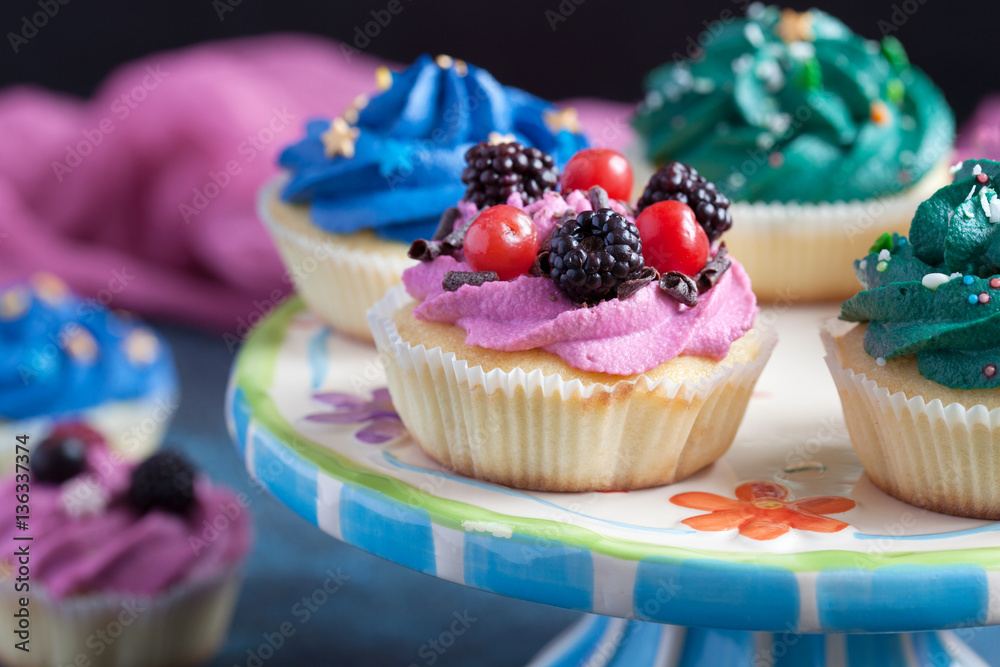 Homemade vanilla cupcakes with multicolor cream cap and berries on a dark blue background.