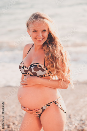 Laughing pregnant girl 20-25 year old holding tummy wearing swimsuit outdoors. Looking away. Happiness. Maternity.