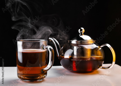 Hot tea in glass teapot and cup with steam on white surface