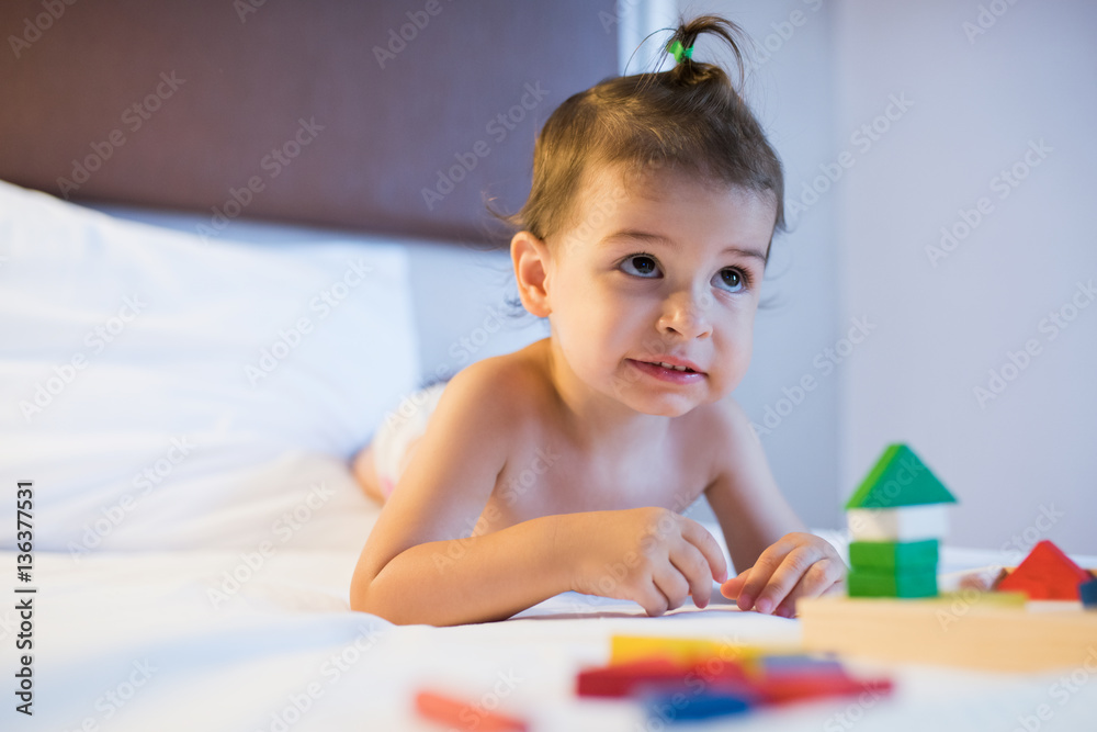 Portrait of toddler beautiful girl lying on the bed in a toy mess playing with colorful construction wooden blocks in the lamplight. Education game for kid in bedroom. Nursery creativity. Portrait of toddler beautiful girl lying on the bed in a toy mess playing with colorful construction wooden blocks in the lamplight. Education game for kid in bedroom. Nursery creativity.