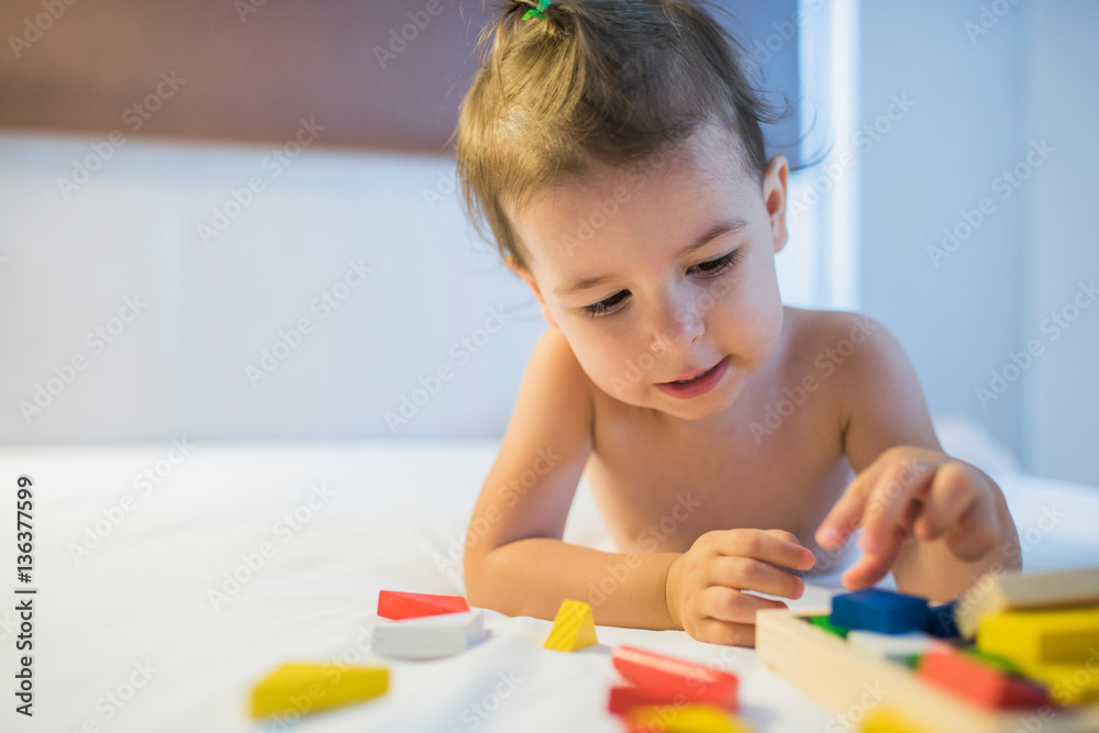 Close-up portrait of beautiful toddler girl lying on the bed in a toy mess playing with colorful construction wooden blocks in the lamplight. Education game for kid in bedroom. Nursery creativity. Close-up portrait of beautiful toddler girl lying on the bed in a toy mess playing with colorful construction wooden blocks in the lamplight. Education game for kid in bedroom. Nursery creativity.