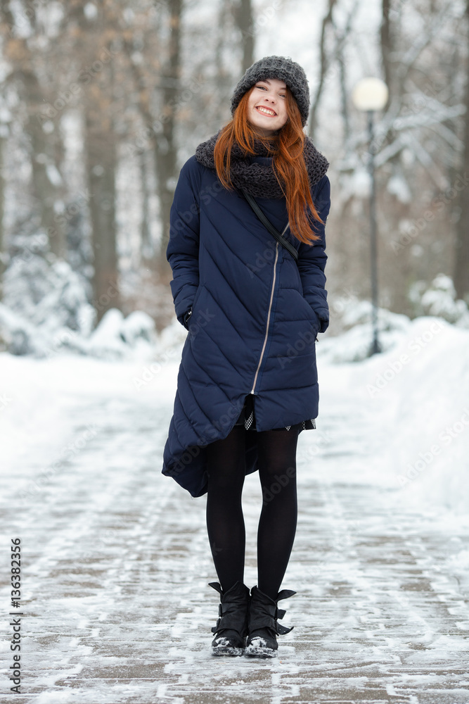 Beautiful winter portrait of young adorable redhead woman in cute knitted hat winter having fun on snowy park path Beautiful winter portrait of young adorable redhead woman in cute knitted hat winter having fun on snowy park path