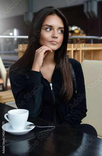 Young beautiful brunette girl in a tracksuit sits in the loft cafe leaning his head on his hand. Woman sitting in cafe thinking. A cup of coffee on the table.