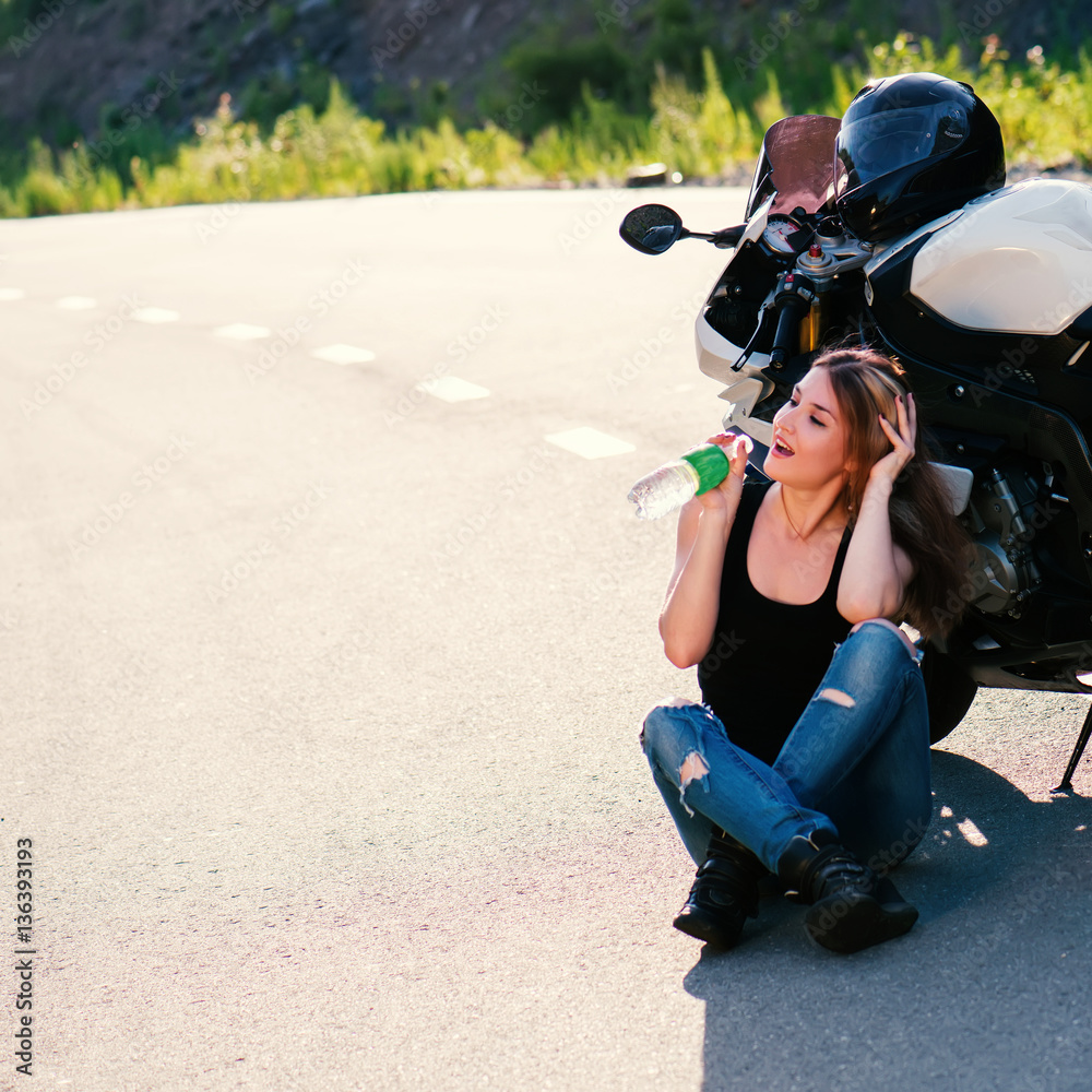 Obraz premium Young beautiful blonde girl in trendy jeans and a black t-shirt sits and drinks water near the modern motorcycle. Outdoor portrait in soft sunlight.