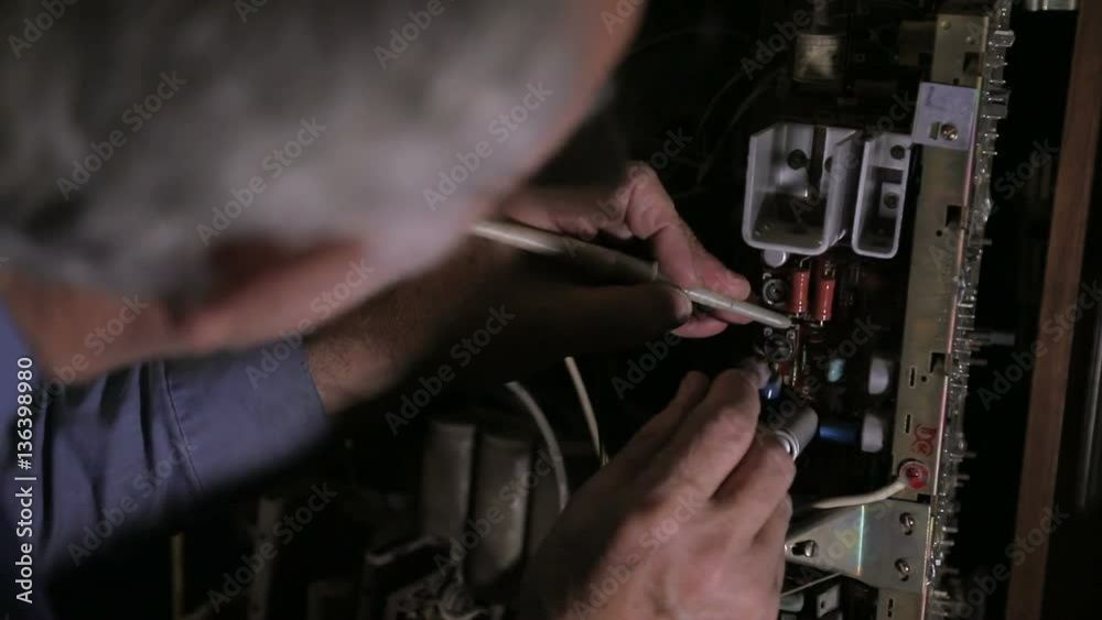 Repairman sits for the work desk with equipment and broken television sets, electrician soldering printed circuit board at the workshop, no color grading, raw video, indoors, concept craftsman. Repairman sits for the work desk with equipment and broken television sets, electrician soldering printed circuit board at the workshop, no color grading, raw video, indoors, concept craftsman.