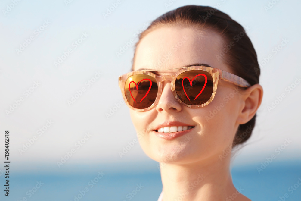 Young beautiful woman wearing sunglasses with hearts on sky background Young beautiful woman wearing sunglasses with hearts on sky background