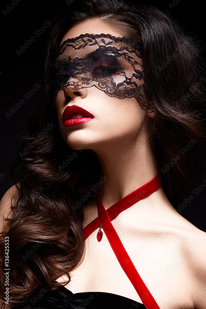 Young girl with a black lace mask on her face and a pendant in the form of heart on the neck. Beautiful model with bright make-up, red lips and curls. The picture on Valentine's Day. Young girl with a black lace mask on her face and a pendant in the form of heart on the neck. Beautiful model with bright make-up, red lips and curls. The picture on Valentine's Day.
