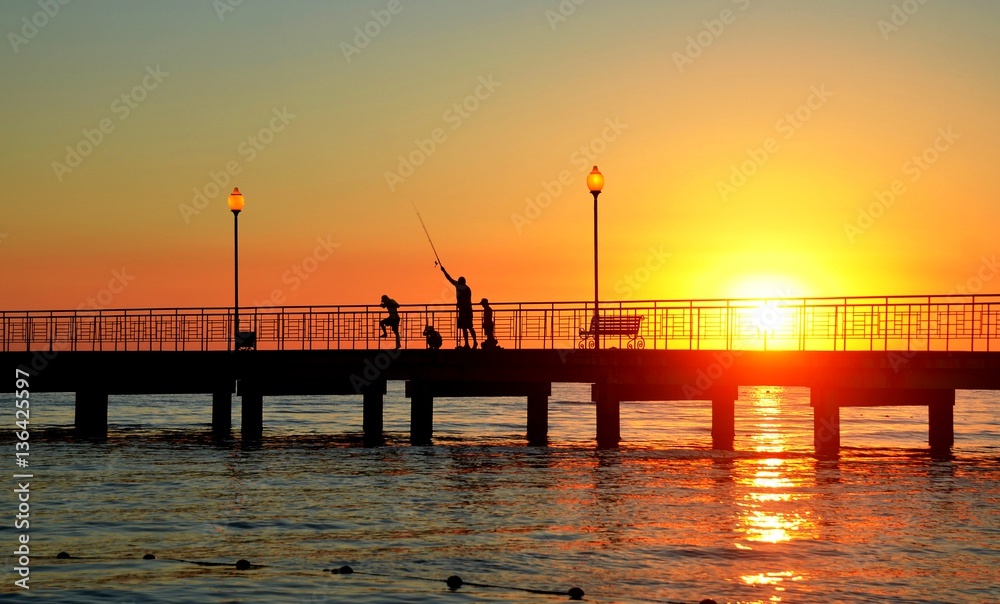 Young father with three young sons, rod catches fish in sea from pier at sunset. Backlight, soft focus, blur effect