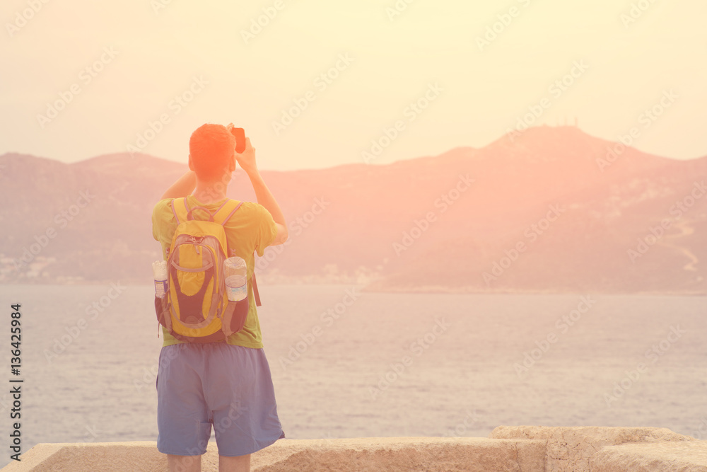 Man with a backpack photographs on the phone the sea and mountains - obrazy, fototapety, plakaty 