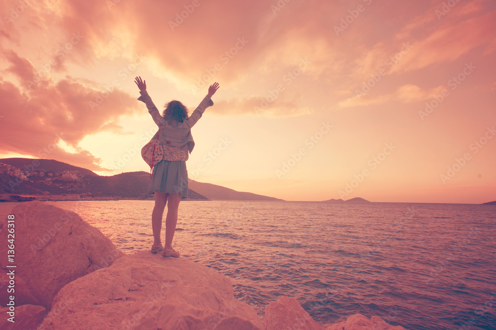 Fototapeta premium Woman with her hands raised standing on a rock near the sea at sunset
