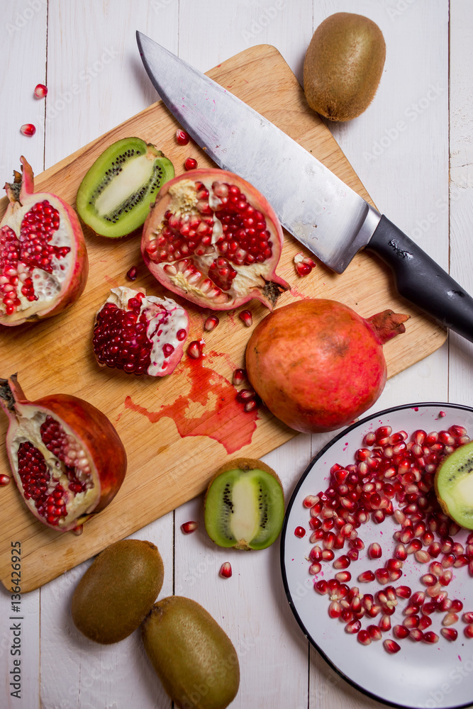 sliced fruit red pomegranate and kiwi on a cutting Board, a plate on white wooden background with knives close-up