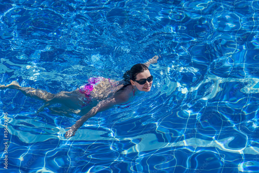 Brunette woman in bikini and sunglasses  swims in the pool