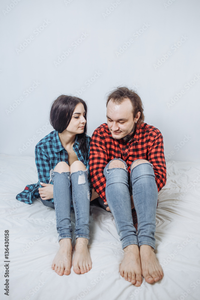 pair of beautiful brunette in a plaid turquoise black shirt and the guy in the red shirt sitting holding hands over the knee and look down on the bed with white blanket, copyspace