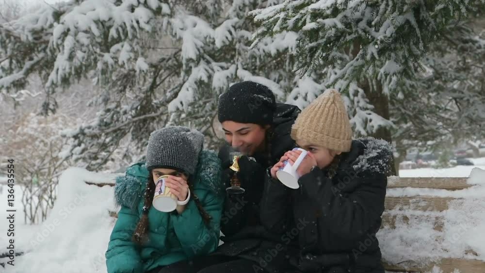 two daughters kiss mom and drinking tea, sitting on a bench on a cold winter day two daughters kiss mom and drinking tea, sitting on a bench on a cold winter day