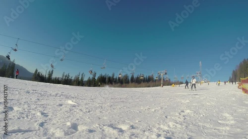 Skiers and snowboarders going downhill on a mountain slope at a ski resort with snowy ski trails in the foreground and mountains, clear blue sky, moving ski lift and evergreen trees in the background