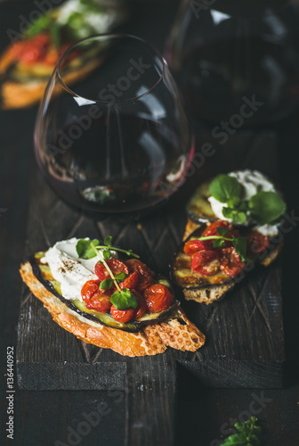 Wine and snack set. Brushetta with roasted eggplant, tomatoes, garlic, cream cheese, arugula and glass of red wine on wooden board over dark background, selective focus. Slow food, party food concept