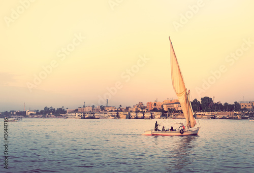 Felucca at sunset - travel on sail vessel on the Nile river, romantic cruise and adventure in Egypt. Traditional egyptian sailboat on horizon. Skyline of Luxor on riverside.