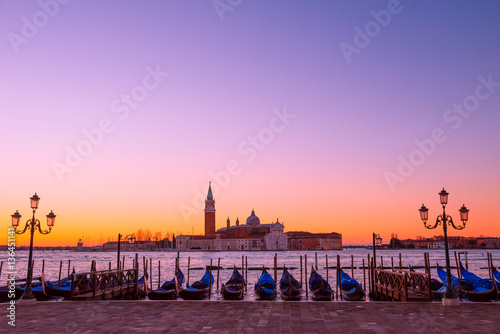 Traditional Italian gondolas moored to the poles in Europe Venice near the city center and Saint Mark square with a background view of the church of San Giorgio Maggiore at sunrise © nevodka.com