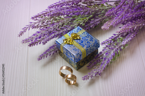 Wedding rings near a blue box near bouquet of artificial purple lavender flowers on the wooden floor