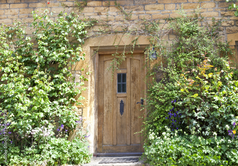 Light brown wooden doors in an old traditional English lime stone cottage surrounded by flowers in the front garden