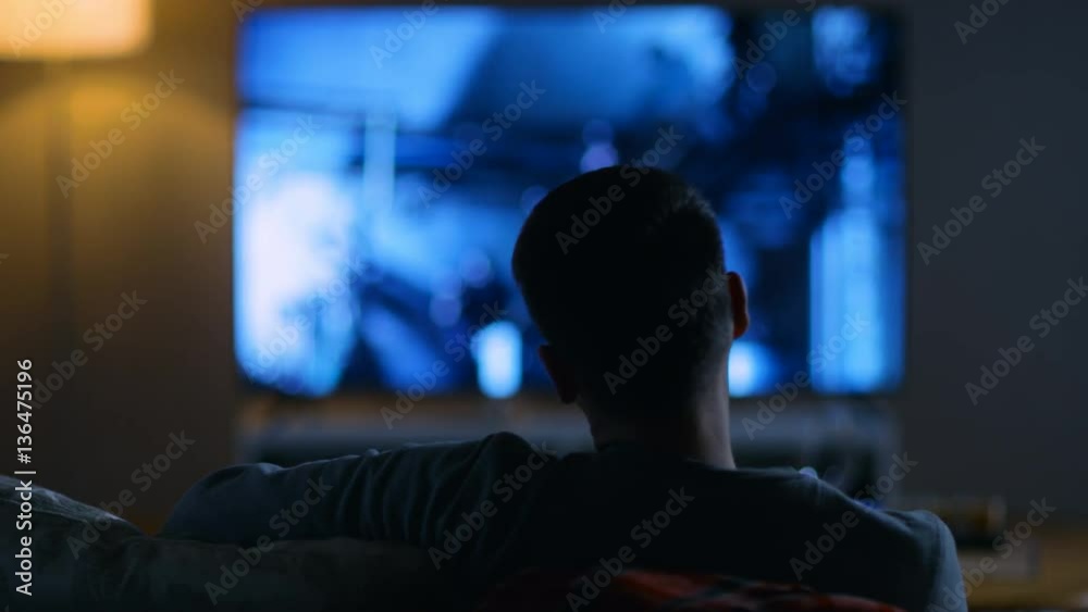 Back View of a Man Sitting on a Couch Watching Movie on His Big Flat Screen TV. Shot on RED EPIC-W 8K Helium Cinema Camera.