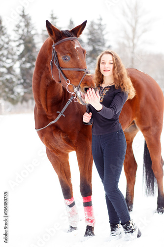 Young rider girl standing with bay horse in winter park
