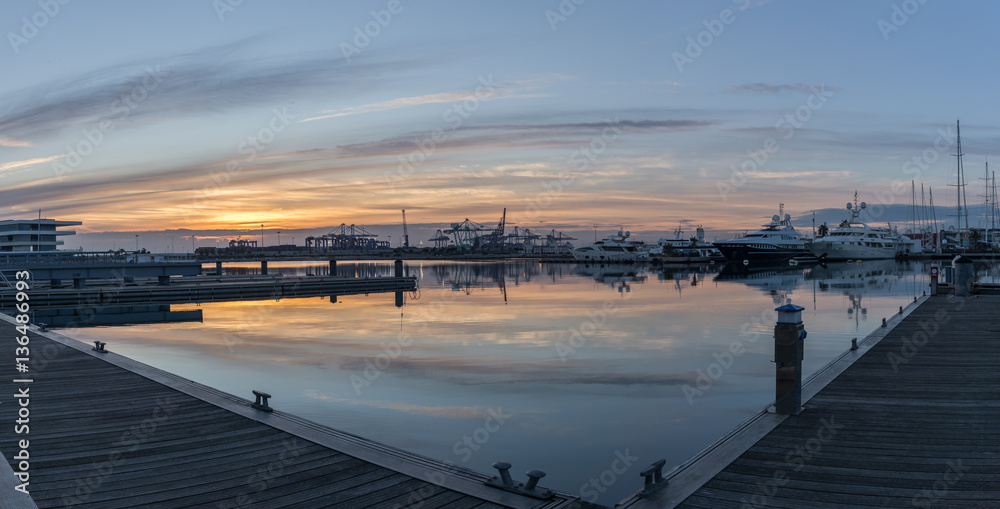 First rays of sun peeking over the horizon. Port twilight in the Valencia harbor cranes working loading transport ships, red and blue skyline reflected in water. gigapan First rays of sun peeking over the horizon. Port twilight in the Valencia harbor cranes working loading transport ships, red and blue skyline reflected in water. gigapan