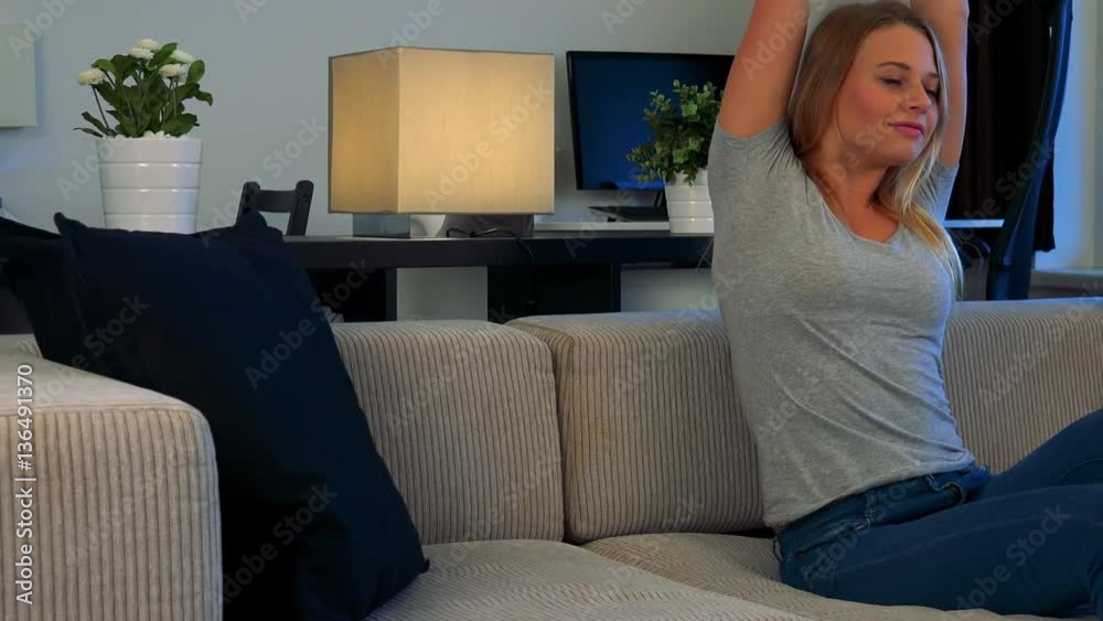 A young, beautiful woman sits on a couch in a living room and stretches