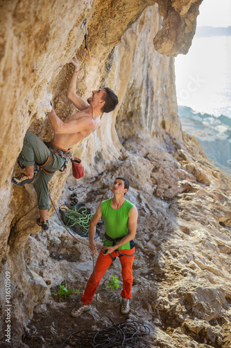 Tableau sur toile Young man starting challenging route on cliff, his partner belay