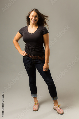 Young mixed race woman wearing a black t shirt and jeans stands with hair blowing and hand on hip while smiling at viewer