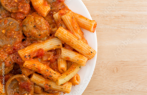 Top close view of rigatoni pasta with meatballs and sausage on a white plate atop a wood table.