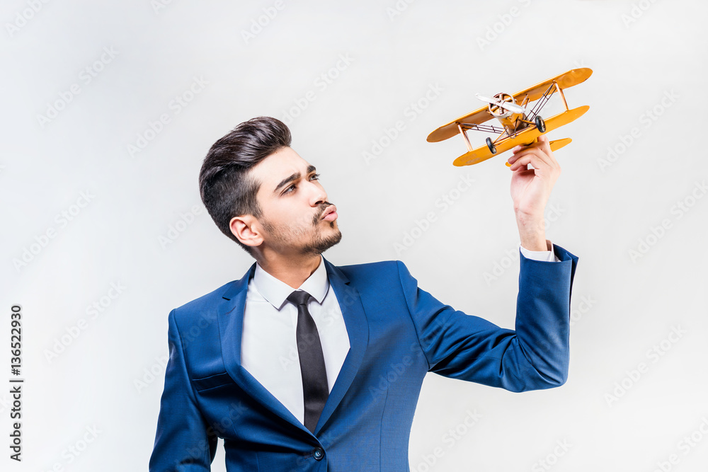 Young attractive man in a blue suit holding a yellow airplane model on a gray background. Isolated