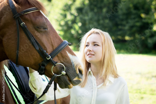 Девушка со светлыми волосами гладит коричневую лошадь и улыбается