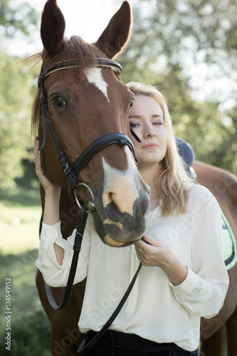 Молодая девушка со светлыми волосами и в белой рубашке стоит рядом с коричневой лошадью 