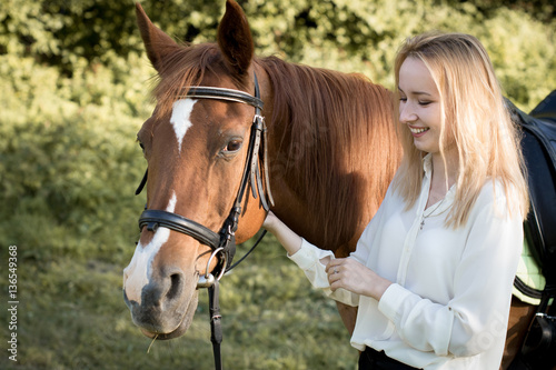 Молодая девушка со светлыми волосами стоит рядом с коричневой лошадью и смеется 